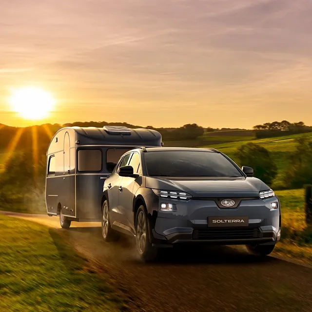 Elektrisk Subaru Solterra trekker campingvogn på landevei i solnedgang over grønne åser.