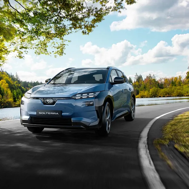 Elbilen kjører langs innsjø i skiftende høstfarger under blå himmel med lette skyer.